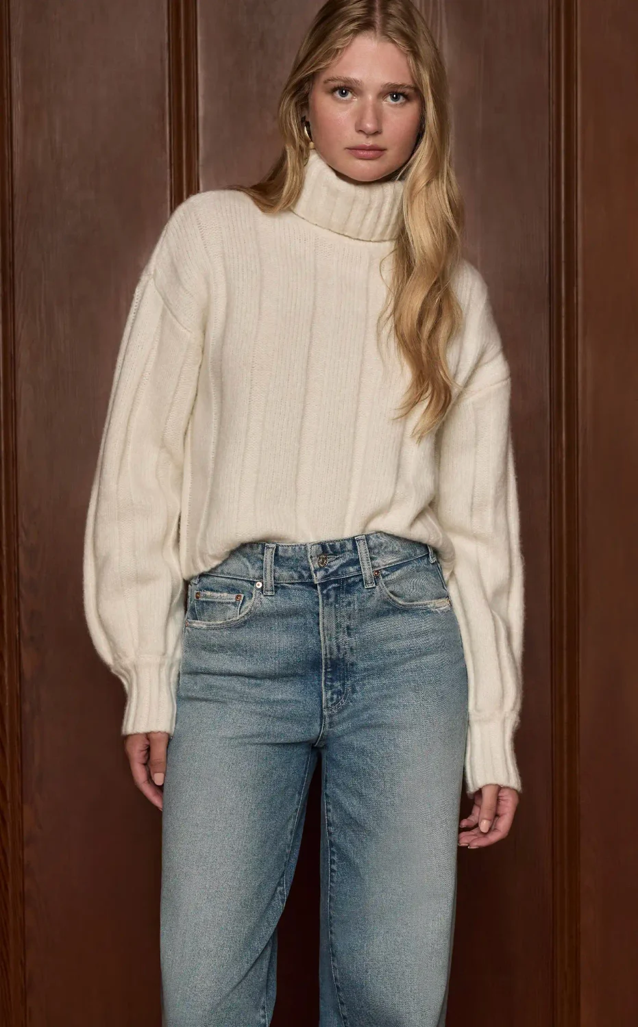 Woman standing wearing an ivory turtle neck sweater and high rise light wash jeans