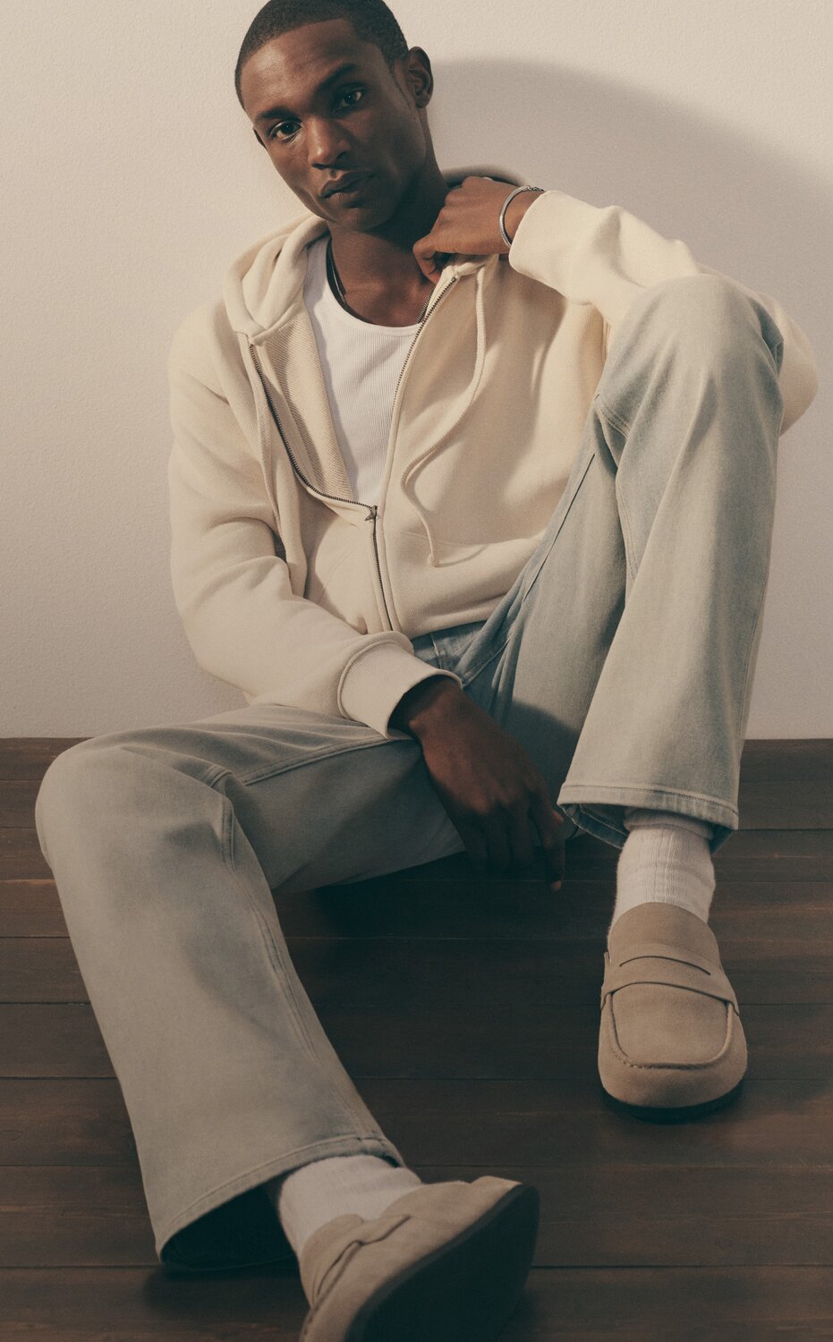 A man sitting on the floor wearing a white t-shirt, a tan zip up hoodie, light blue denim jeans and tan loafers. 