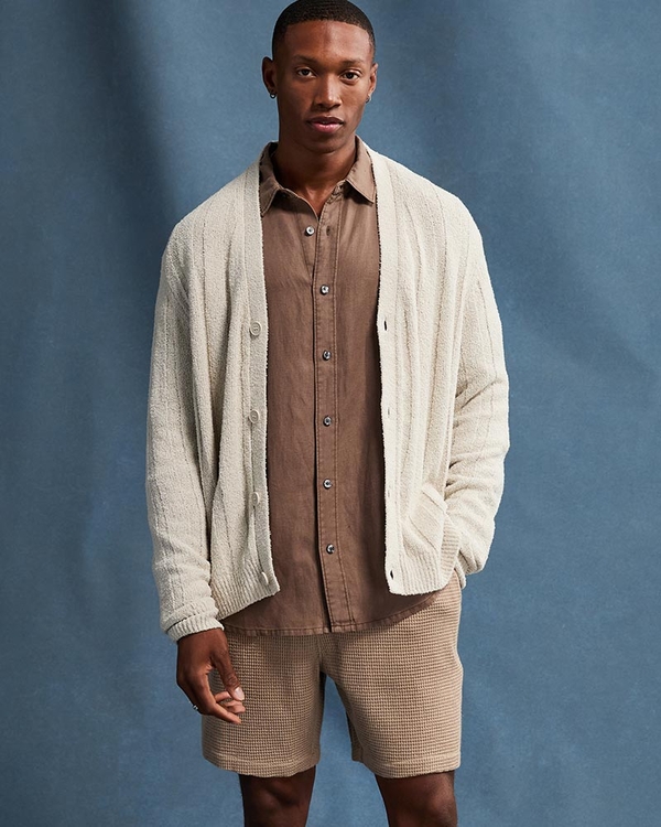 Man wearing a cream cardigan over a brown button up shirt and light brown shorts