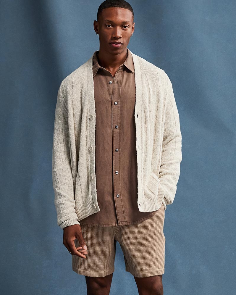 Man wearing a cream cardigan over a brown button up shirt and light brown shorts