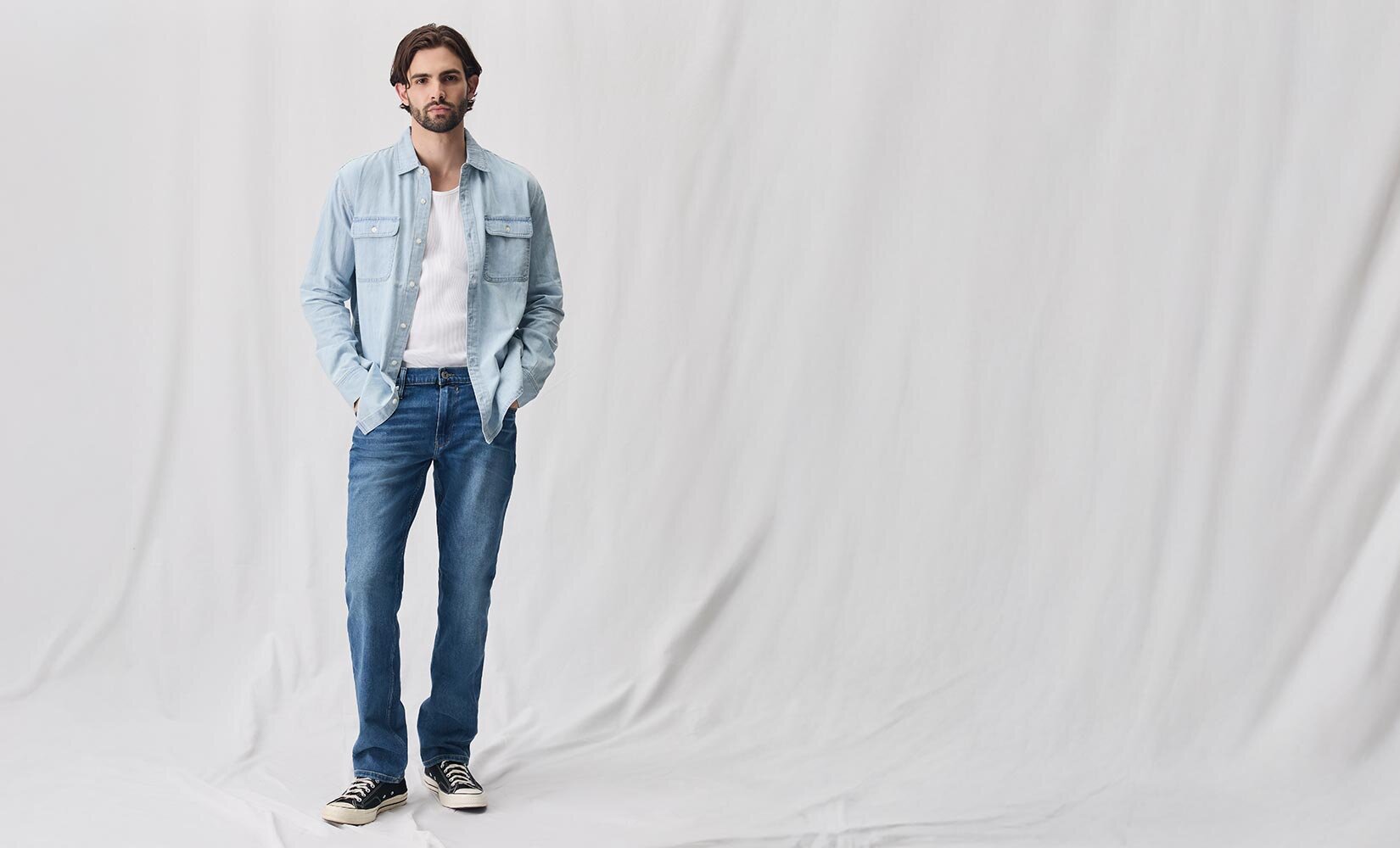 Man wearing a light wash denim shirt with a white tank top underneath, medium wash jeans and black sneakers