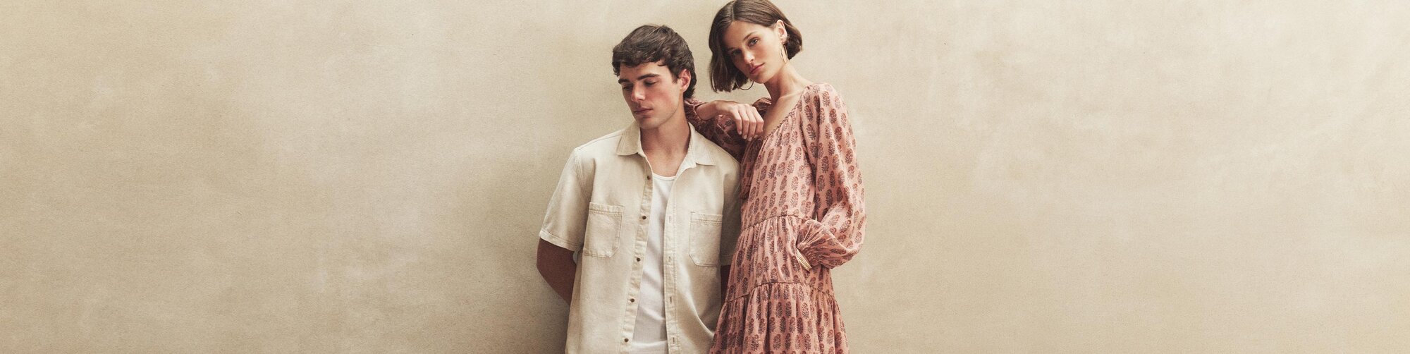 Man wearing a biege short sleeve button up denim shirt with white ribbed tank standing next to a woman wearing a pink long sleeve mini-dress with embroidered pattern detail.