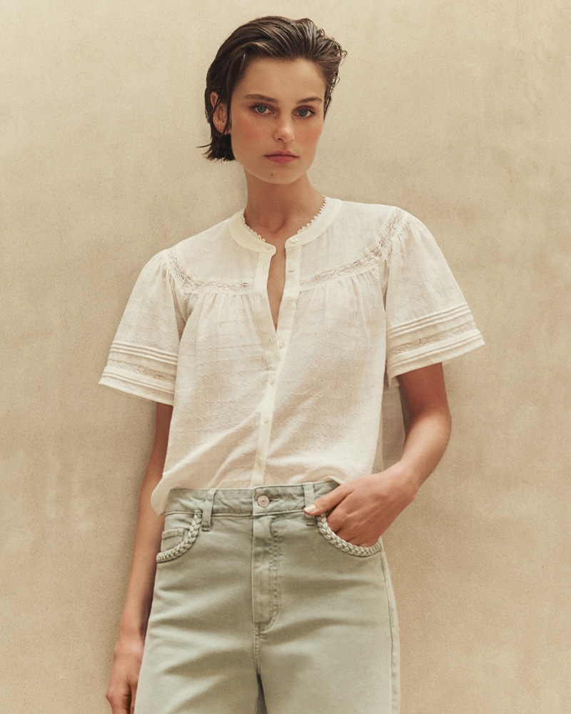Model wearing a white short-sleeve blouse with lace detailing, paired with high-waisted green pants, against a neutral backdrop.