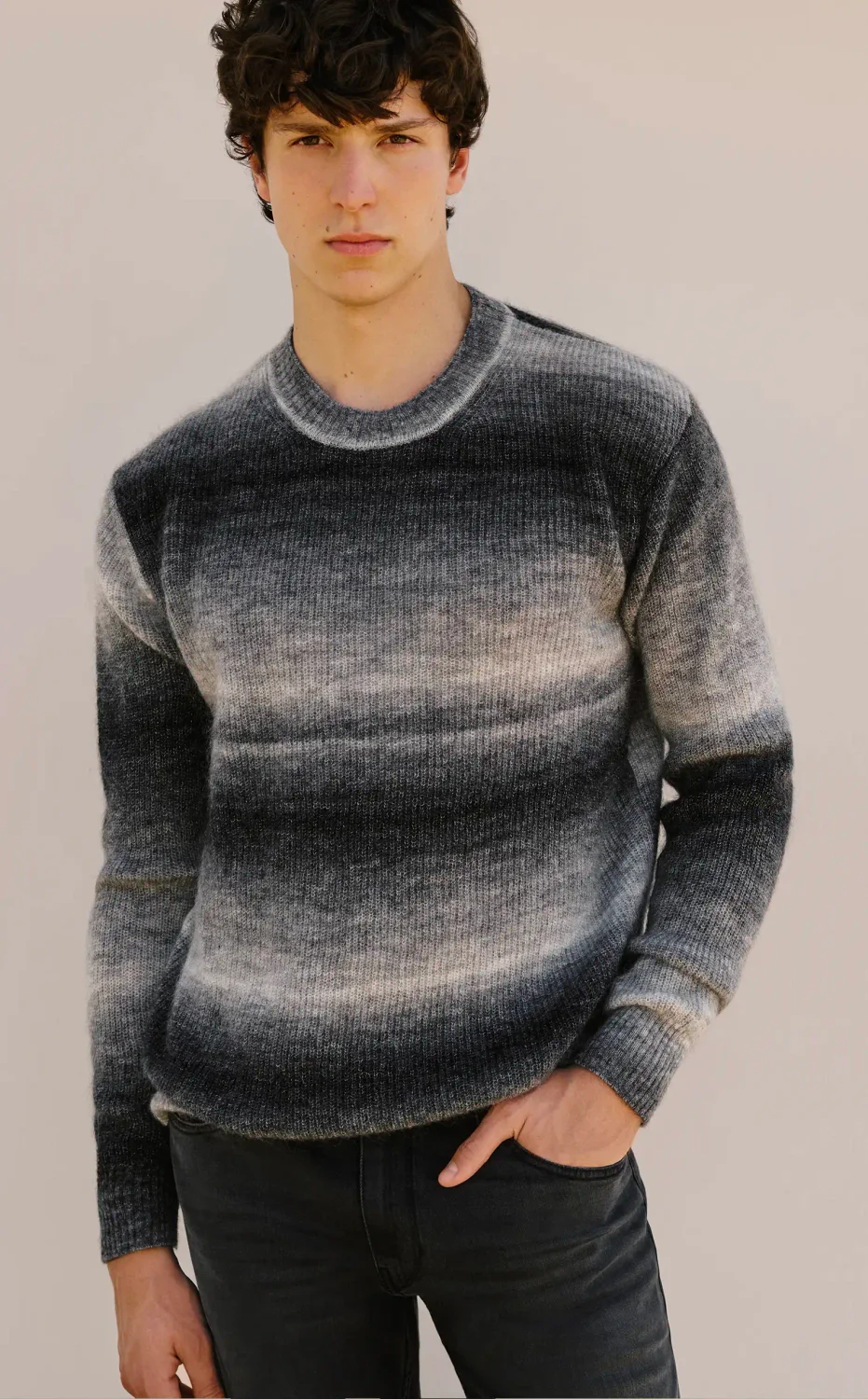 Man standing wearing a grey and black gradient striped sweater and dark wash jeans