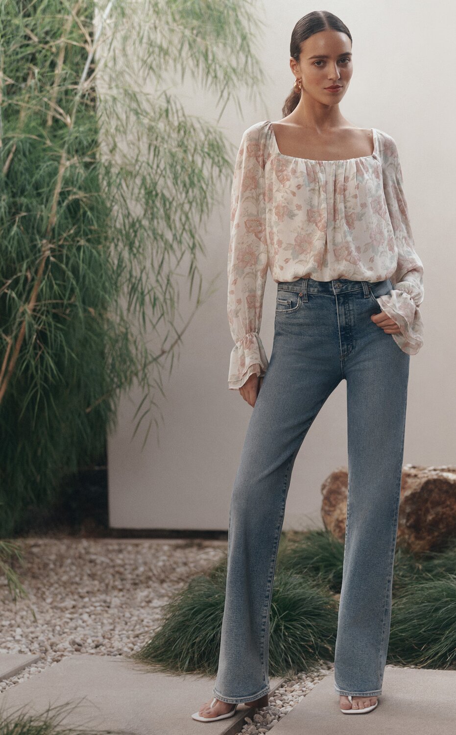 Woman wearing a long-sleeve floral blouse and light-wash high-rise bootcut jeans standing outdoors against a cream wall and green foliage.