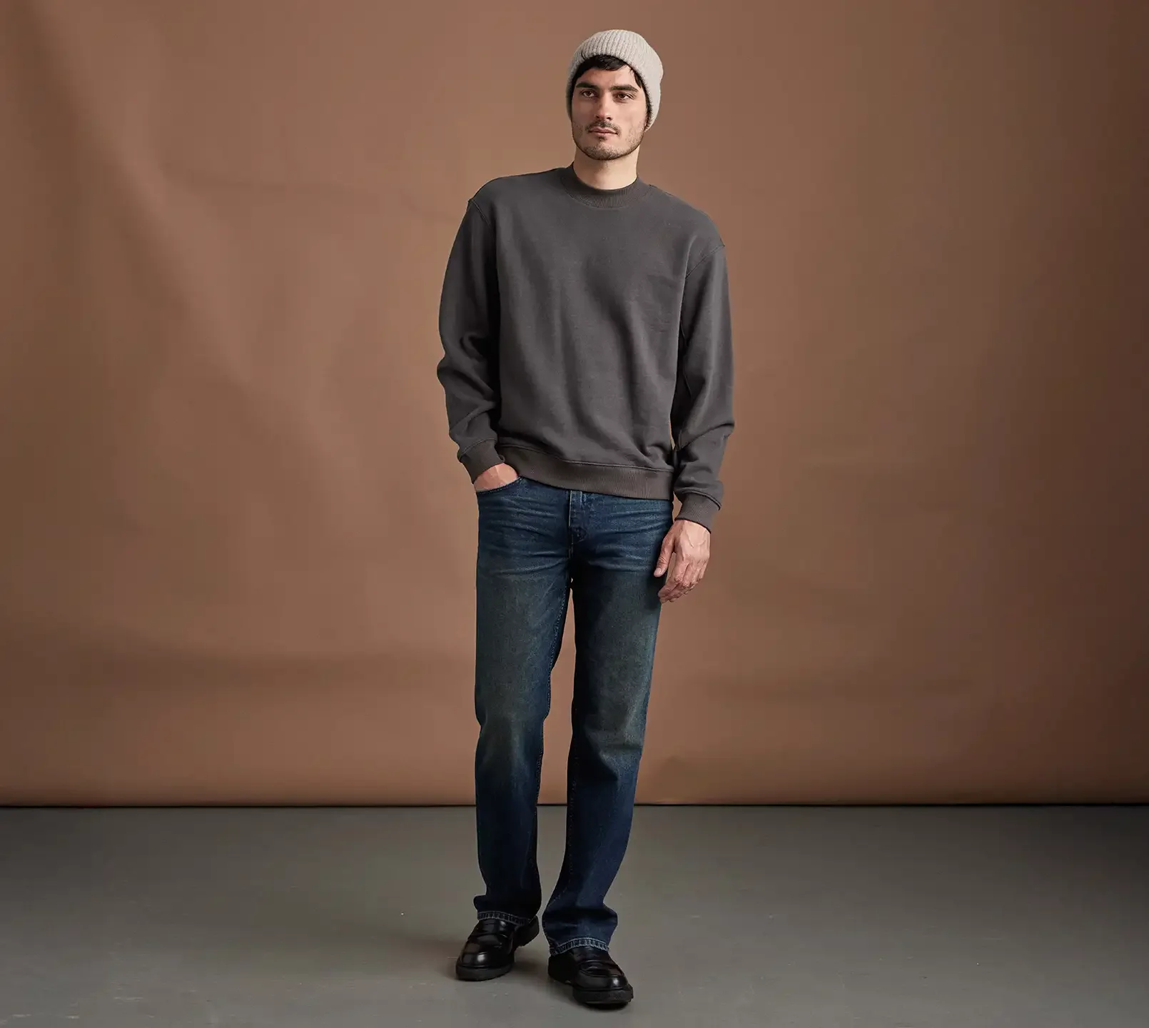 Man standing wearing a sand beanie, a grey crewneck pullover and relaxed dark wash jeans