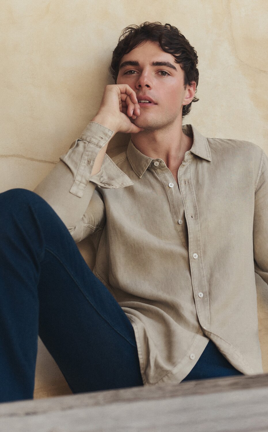 Man lounging in a taupe long-sleeve button-front shirt and dark wash jeans.