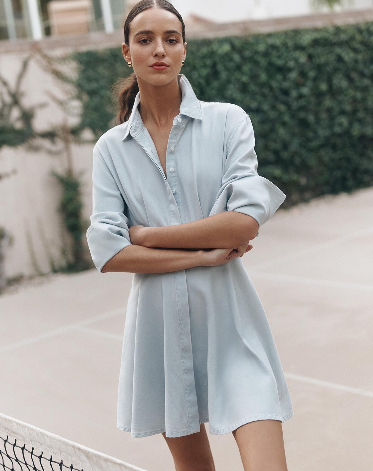 Woman wearing a light blue shirt dress with rolled sleeves, standing outdoors with arms crossed.