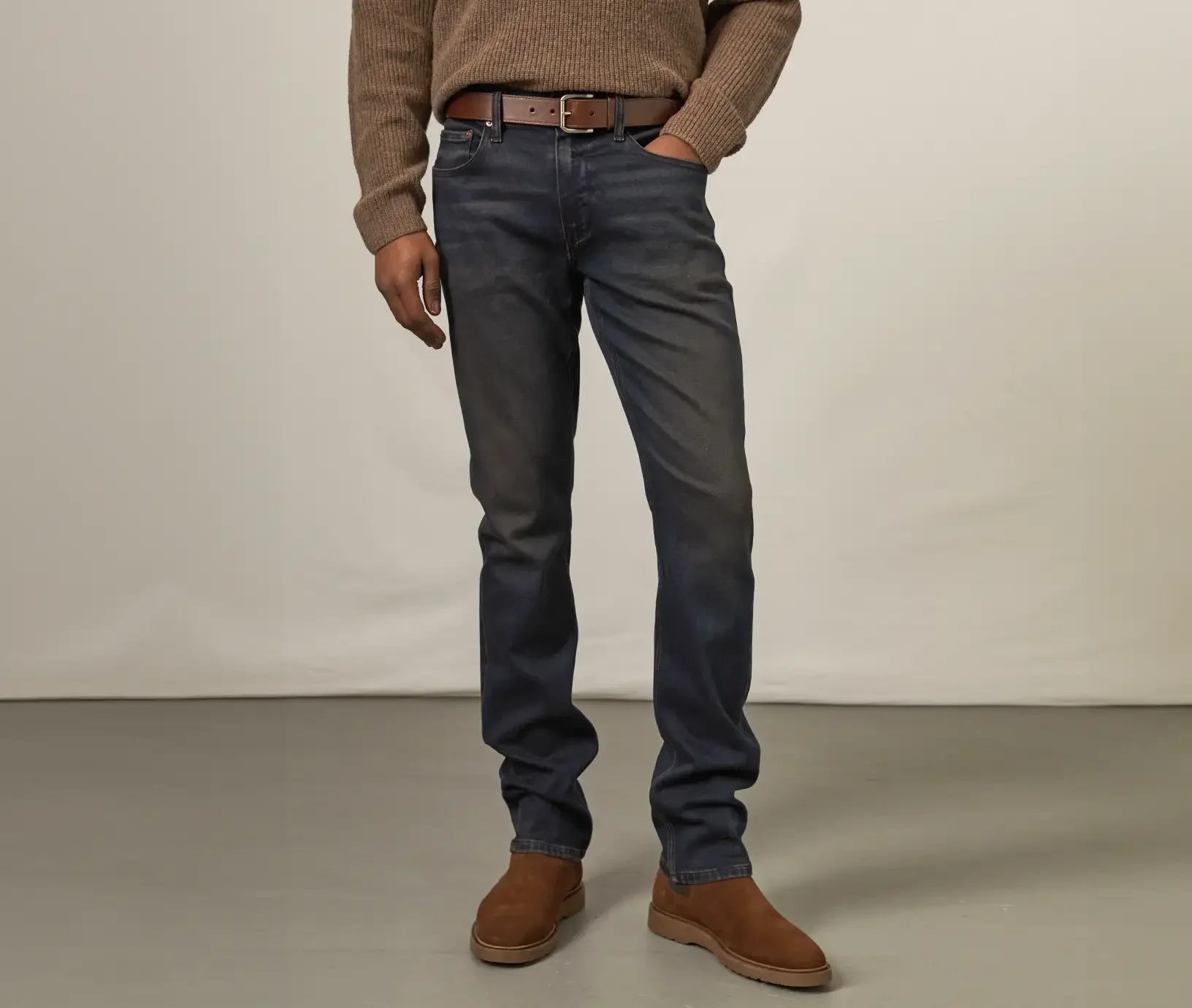 Man standing wearing dark wash jeans, a brown belt, brown boots and a brown sweater