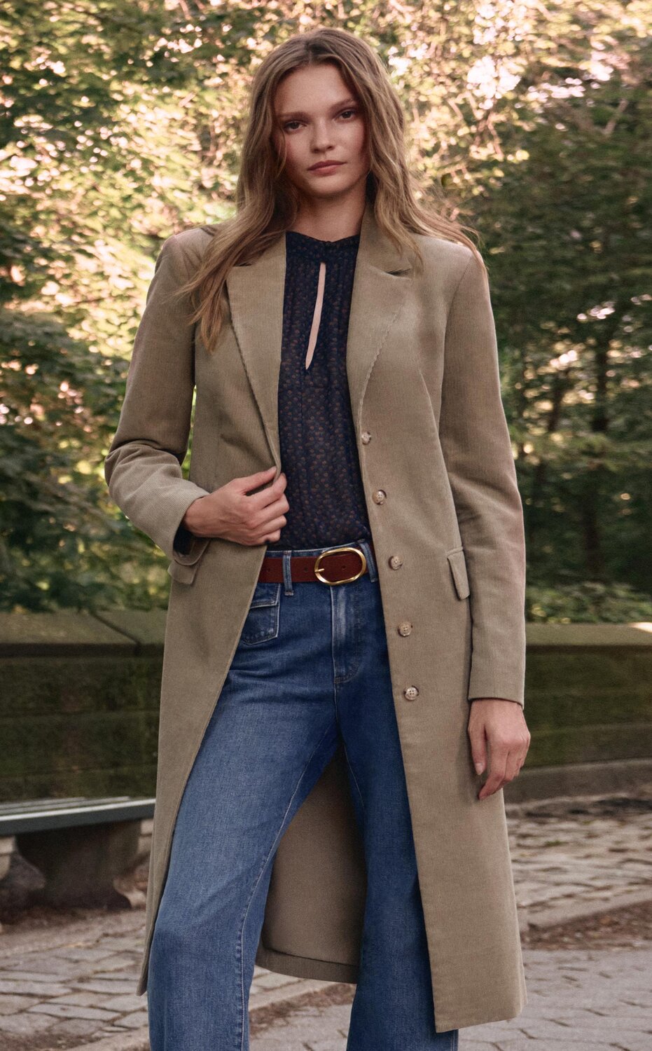 Woman wearing a long olive green corduroy coat over a blue blouse, wide leg jeans, a brown belt and brown boots