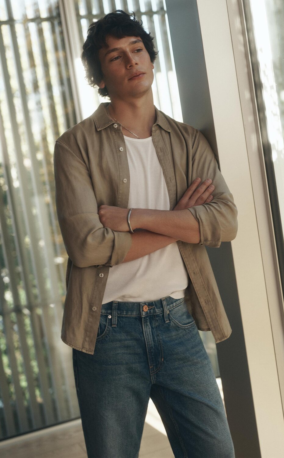 Man wearing a light beige button-down shirt over a white tee and blue jeans leaning against a window.