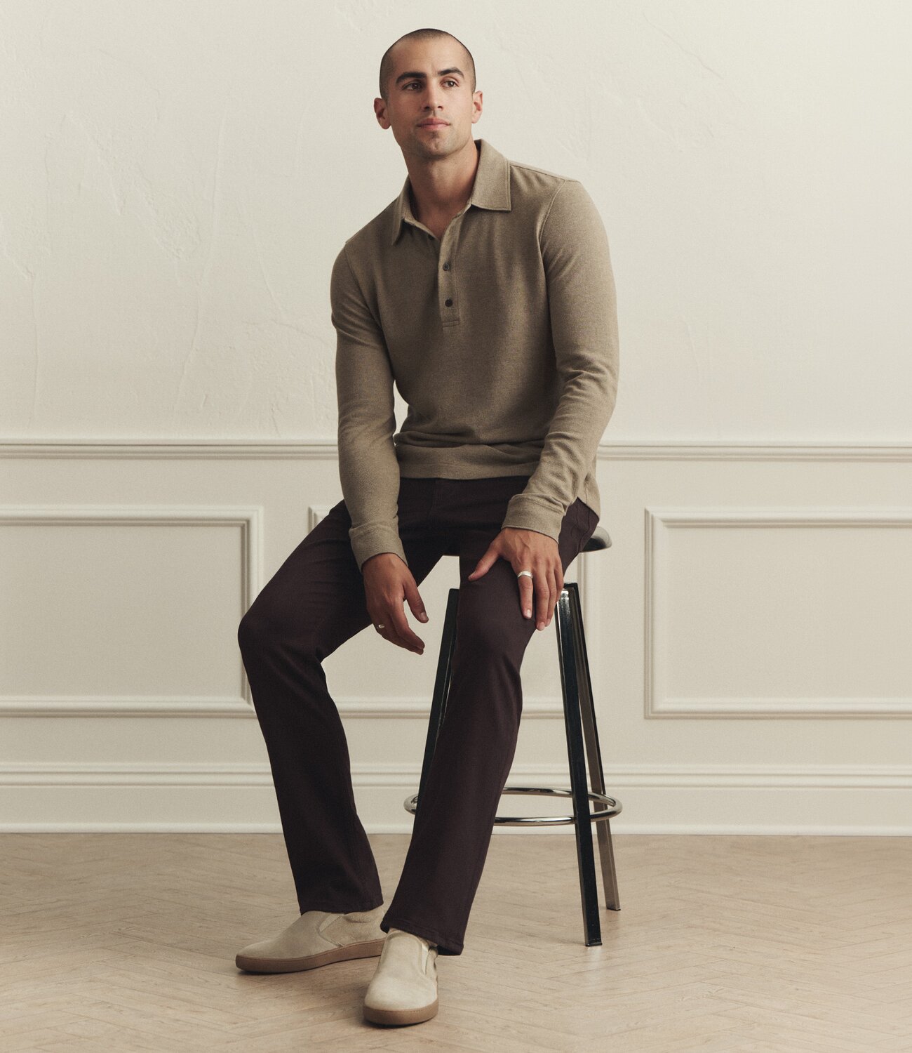 Man wearing a muted green long-sleeved polo knit blouse with a pair of dark brown straight-leg pants and beige suede slip-on sneakers, lounging on a stool against a cream wall with trimming and beige hardwood floors.