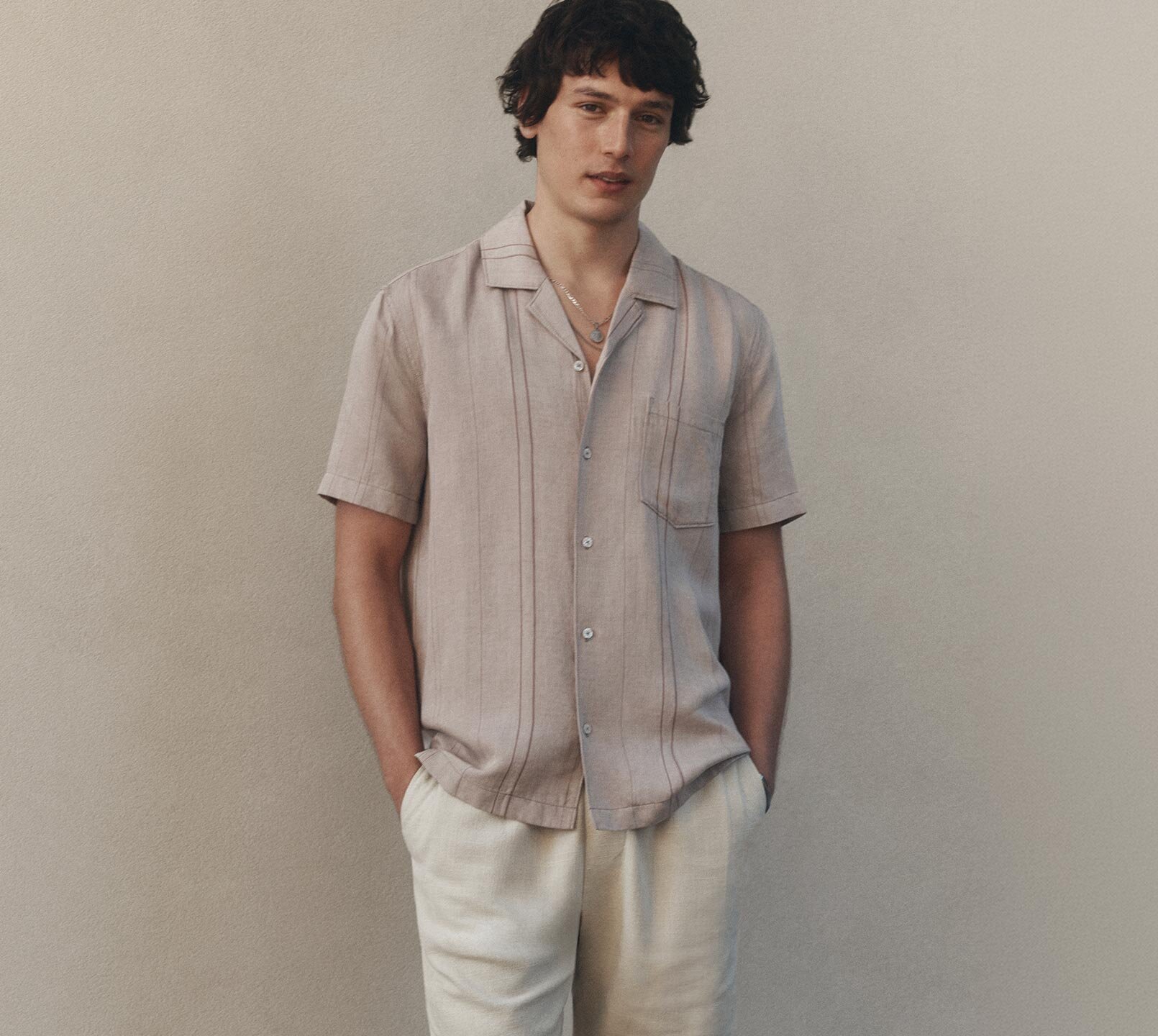 Man wearing a light pink short sleeve button up shirt with vertical broken stripe detail paired with light cream linen shorts.