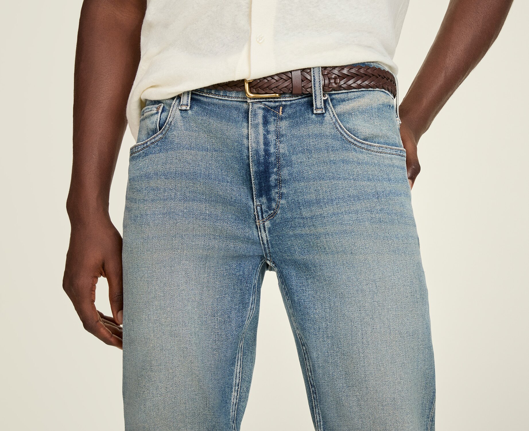Close-up of a model wearing light-wash slim-fit jeans paired with a cream short-sleeve shirt and a brown woven leather belt, highlighting the denim fit and wash.