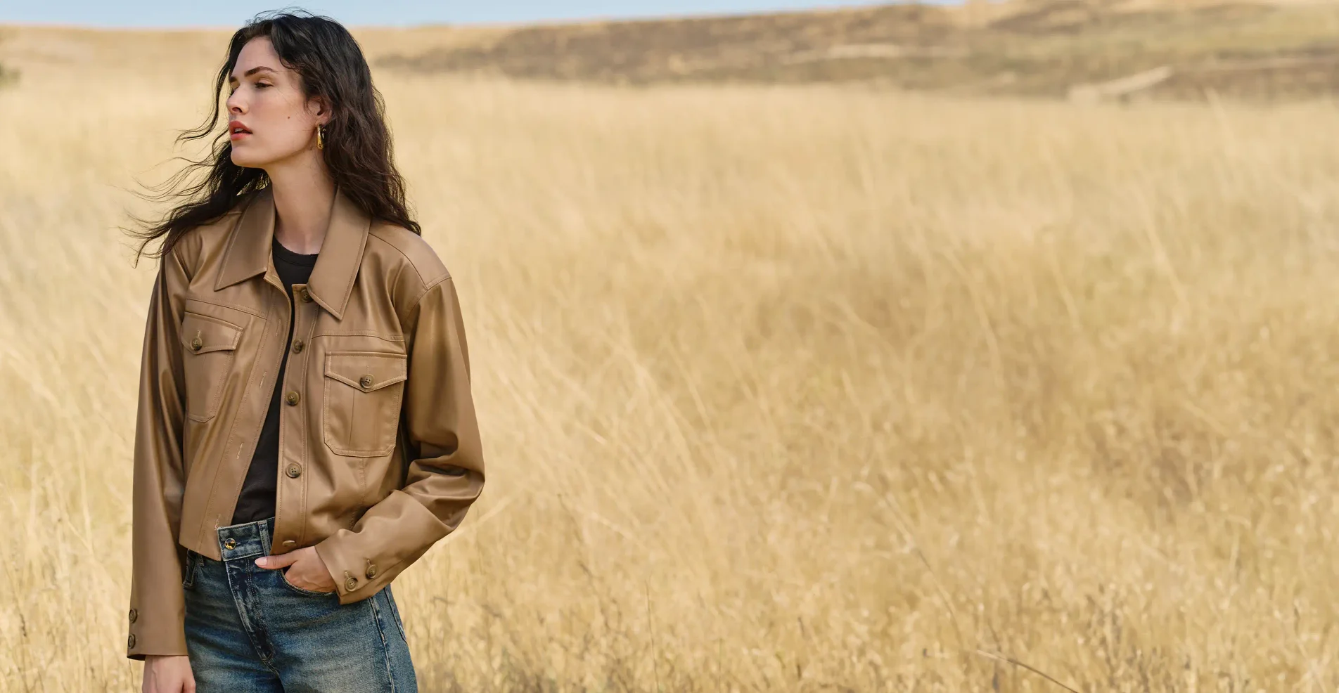 Woman standing outside in a field wearing a light brown faux leather button up jacket, a black top and dark wash jeans