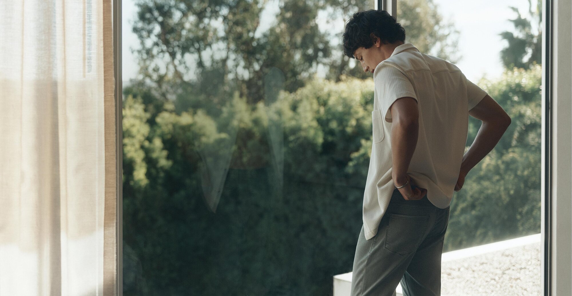 Man wearing a tan button-down shirt over a white T-shirt and blue jeans looking out a window.