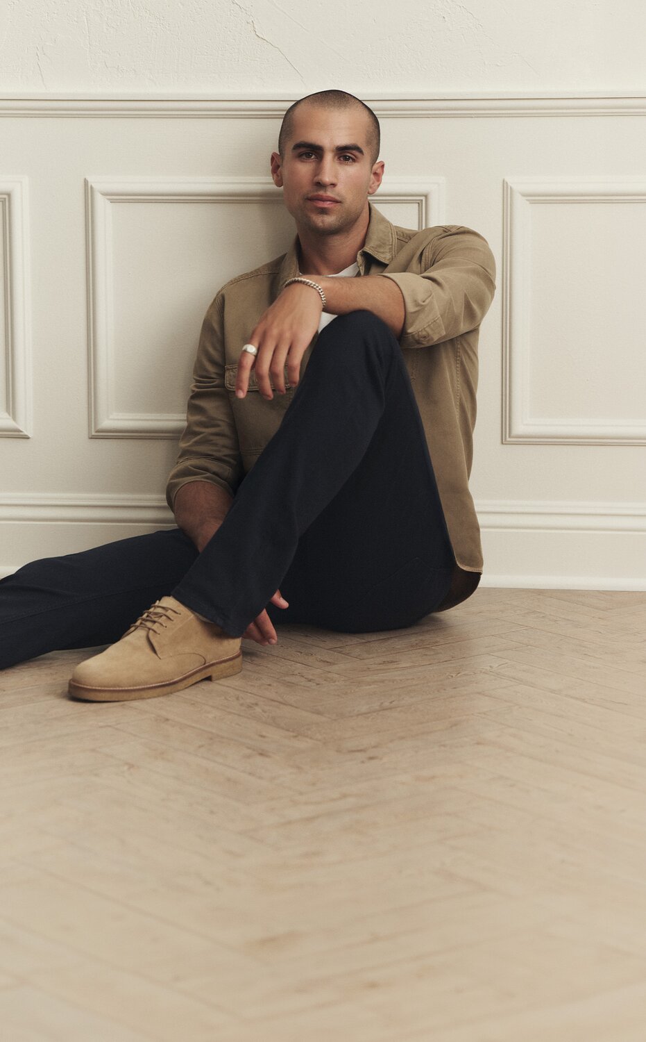Male model wearing dark wash PAIGE jeans styled with a tan button-down shirt and brown suede shoes.