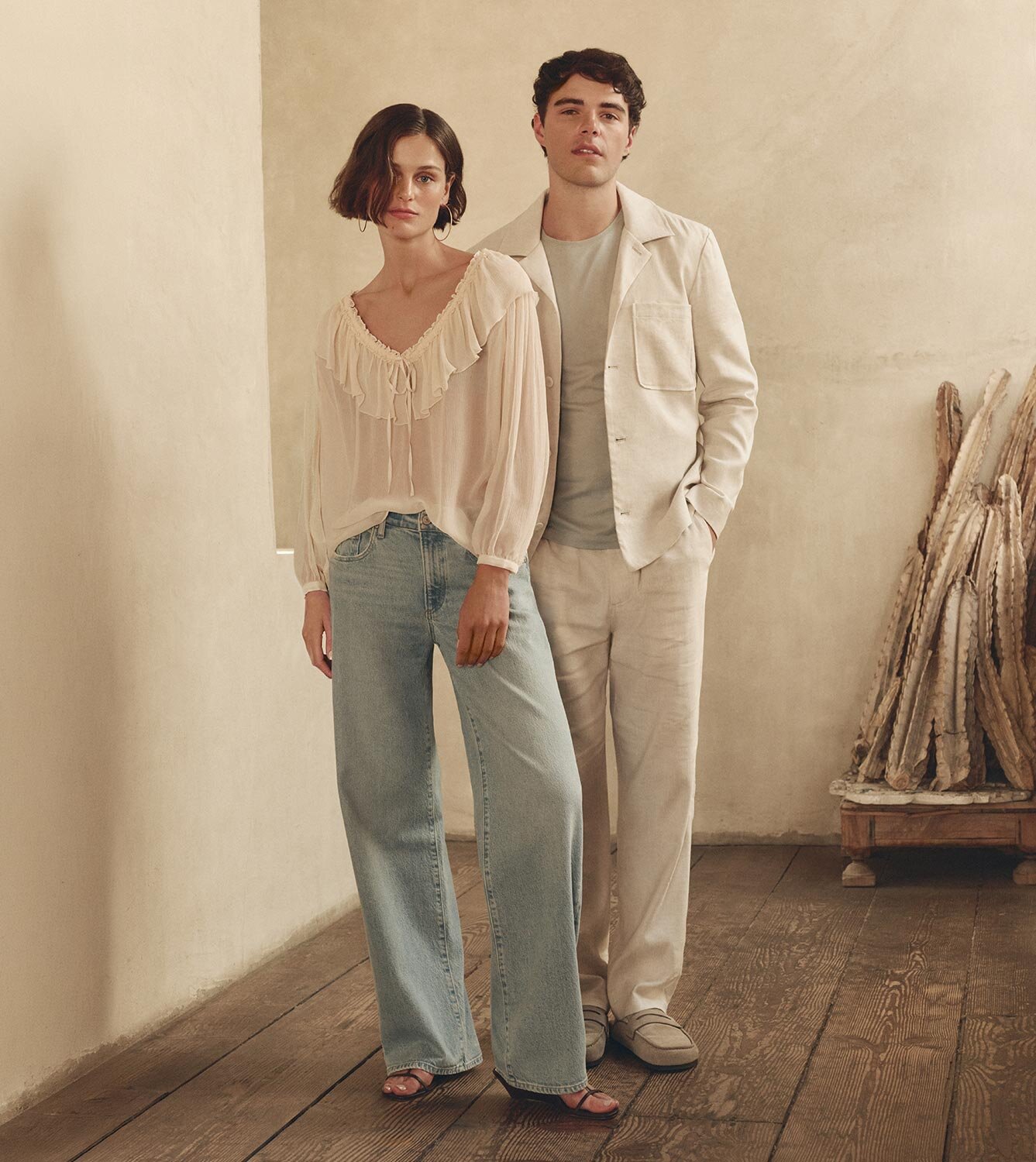 Image of a woman wearing a very light cream colored blouse paired with light washed blue denim jeans and brown leather strappy sandals standing next to a man wearing a light cream colored linen jacket and matching pants paired with a light blue t-shirt and light tan suede loafers.