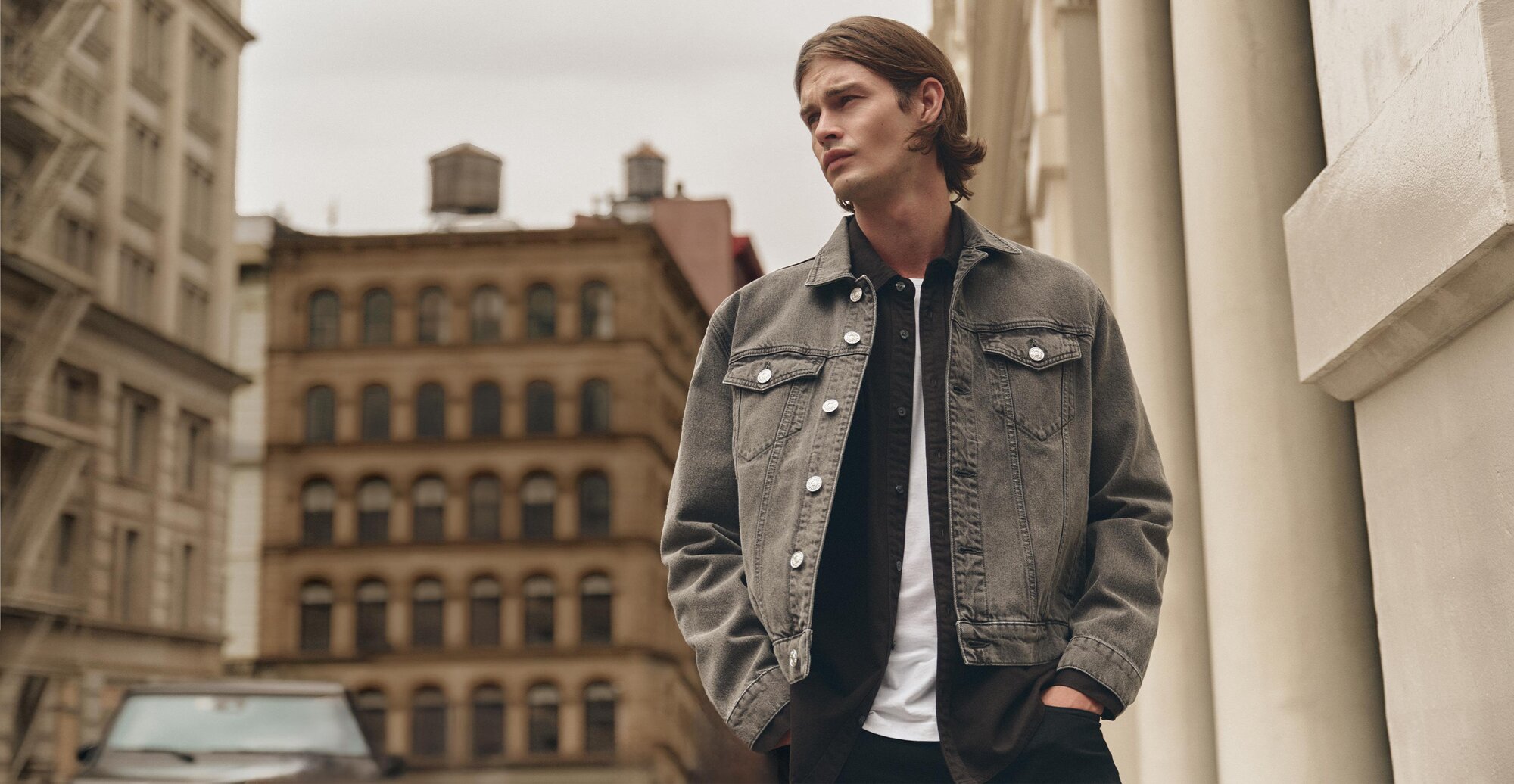 Man wearing a dusty brown wash denim jacket over a dark brown button up shirt, white tee, and dark wash jeans