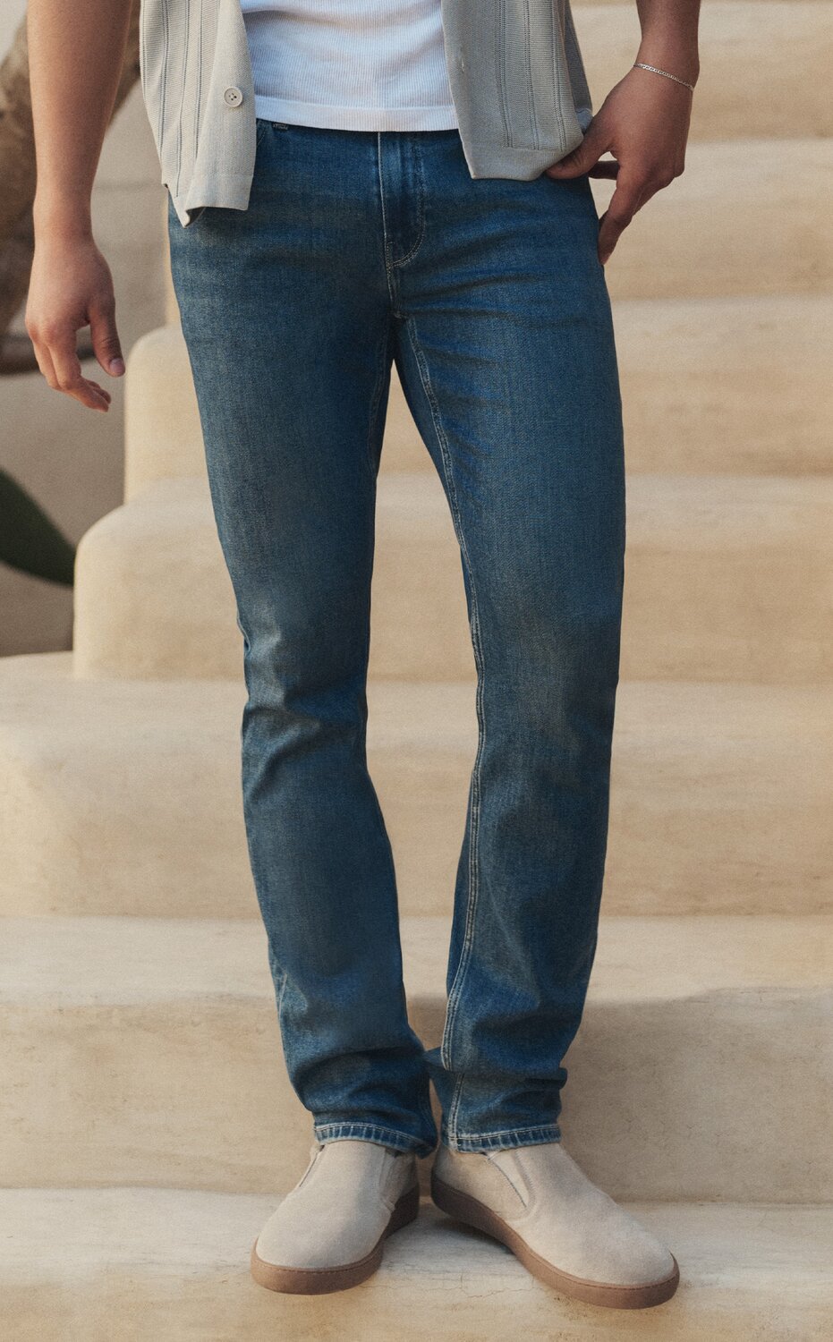 Male model wearing a short-sleeve button-up shirt over a white tee with blue jeans, standing on a sunlit stone staircase.