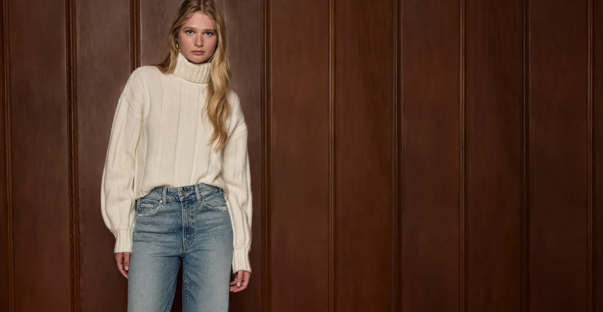 Woman standing wearing an ivory turtle neck sweater and high waisted light wash jeans