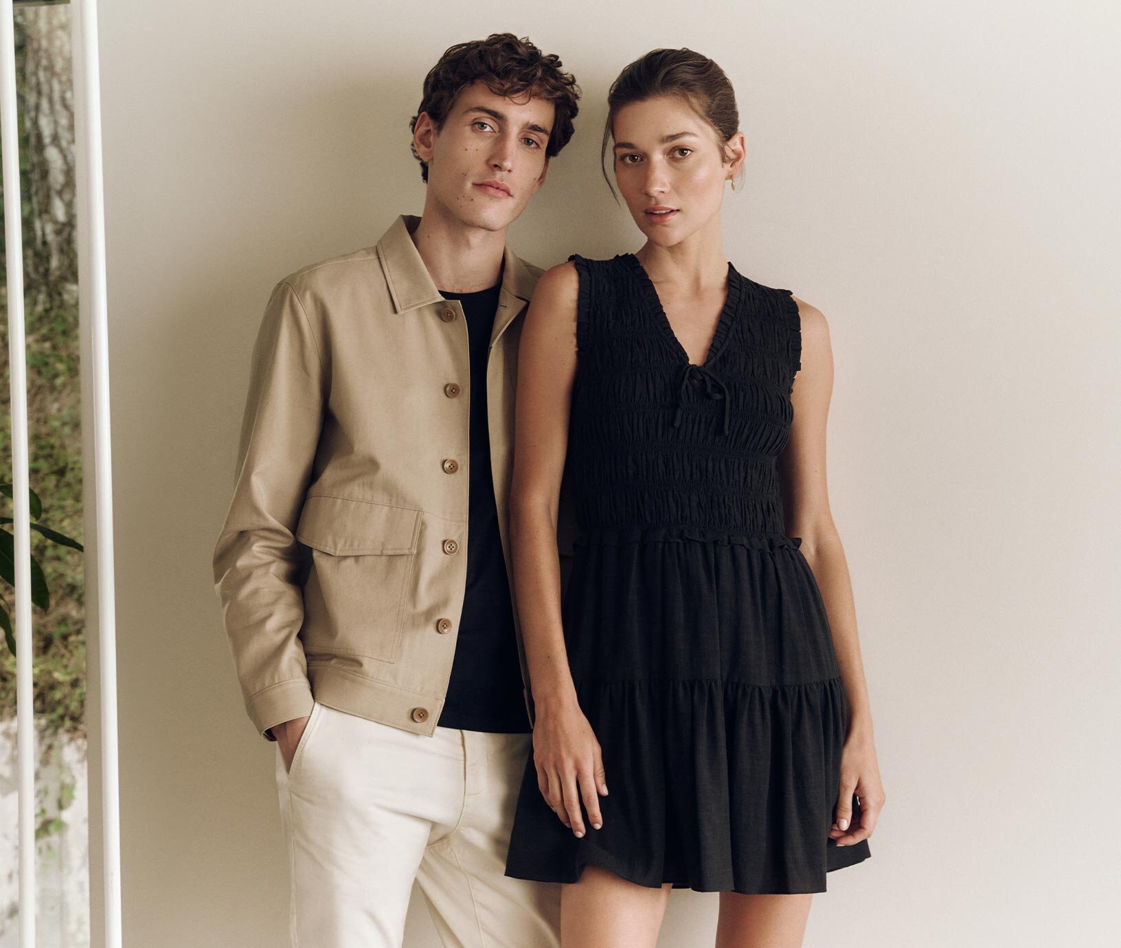 Male model wearing a beige cotton twill cropped trench, ecru chinos and black t-shirt next to female model wearing a black mini dress in a textured cotton all by PAIGE