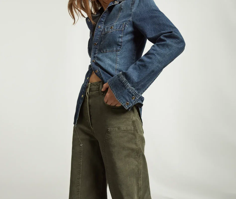 Woman standing wearing a dark wash denim button up shirt and dark green utility pants