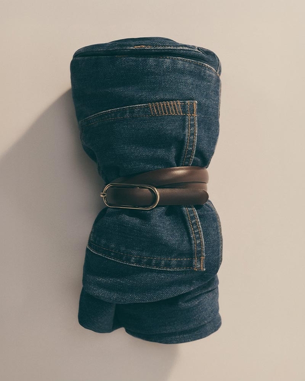 A man wearing a light grey short sleeve t-shirt and dark blue denim jeans with his hands in his pockets, leaning against a white wall