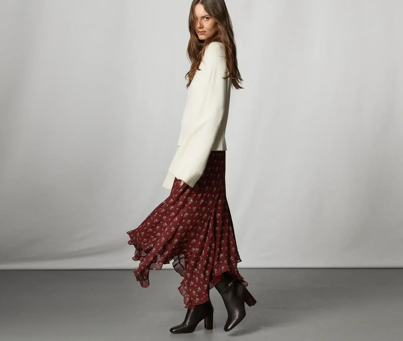 Woman standing wearing a white sweater, burgundy floral midi skirt and tall brown boots