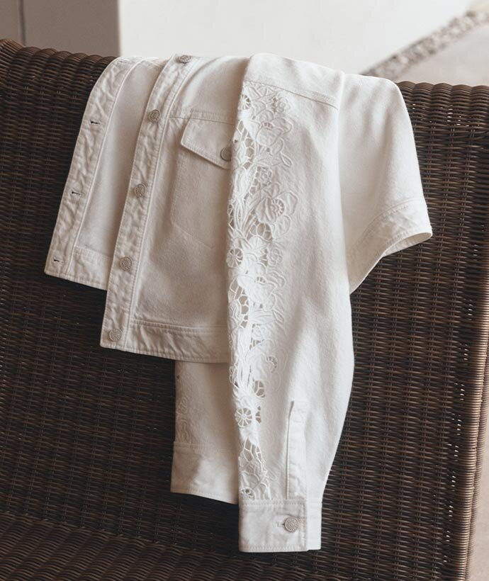 A white denim jacket with floral lace sleeve detail laid over a brown wicker chair.