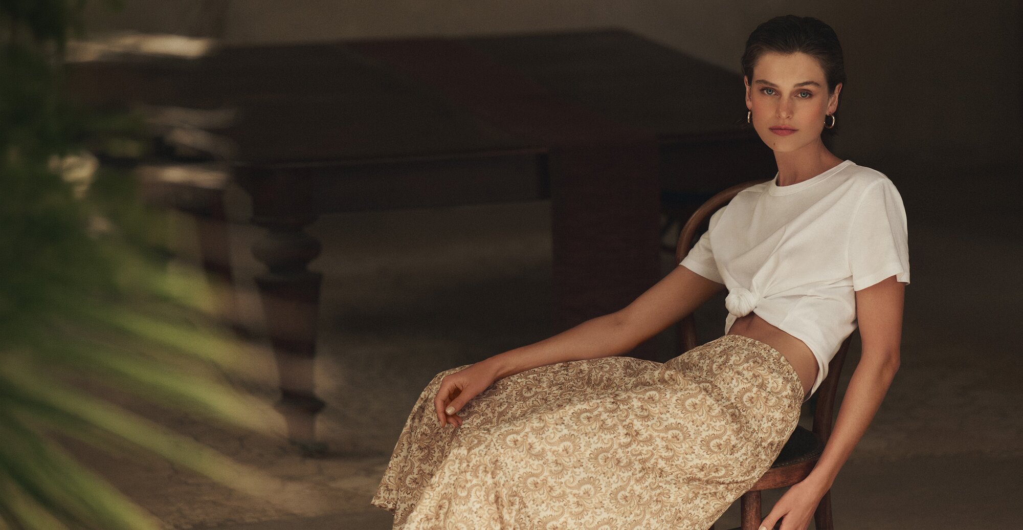 Woman wearing a white tee and a patterned midi-length skirt, lounging on a chair in an outdoor area.