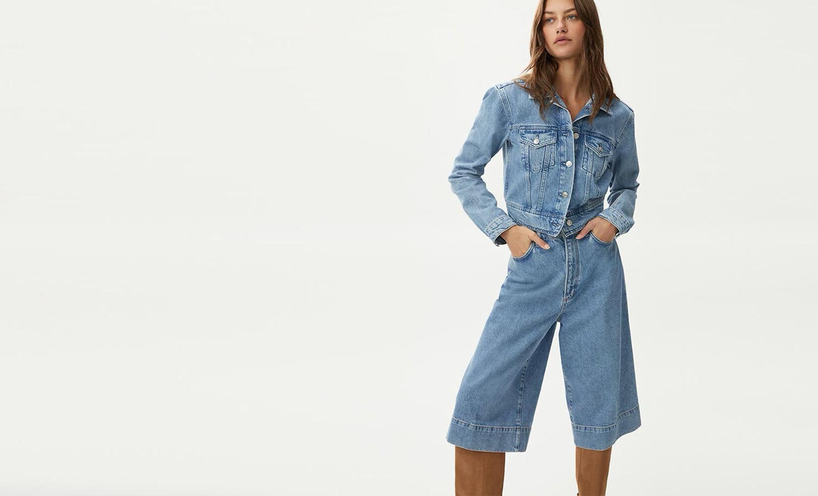 Woman wearing a light wash denim on denim jacket & gaucho set with tall brown suede boots
