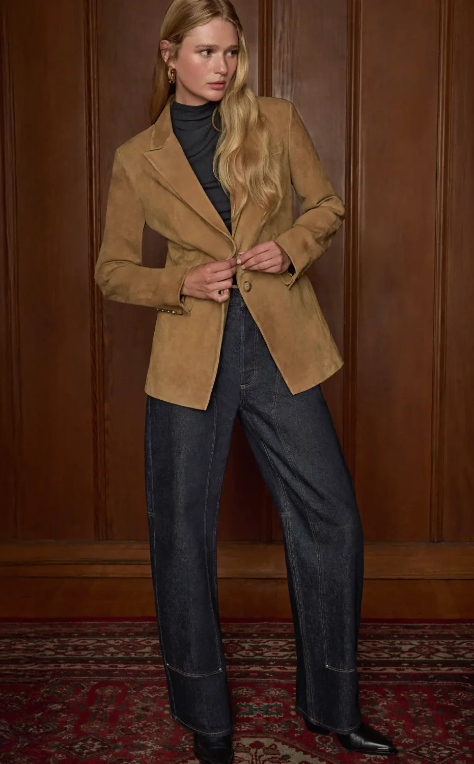 Woman standing wearing a tan suede blazer, a black top, and dark wash workwear jeans