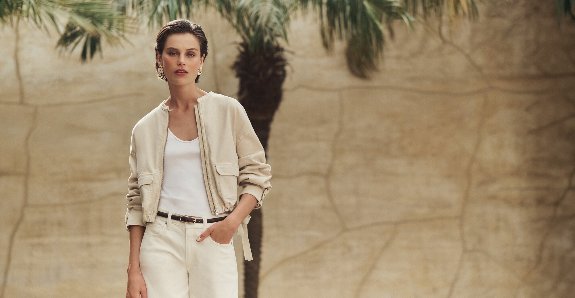 Female model wearing a cream-hued jacket with an adjustable tie waist and cargo-style pockets over a white scoop-neck tank and a pair of cream high-waist jeans fastened with a thin dark brown leather belt. She poses in front of a sunlit stone beige wall and trees.
