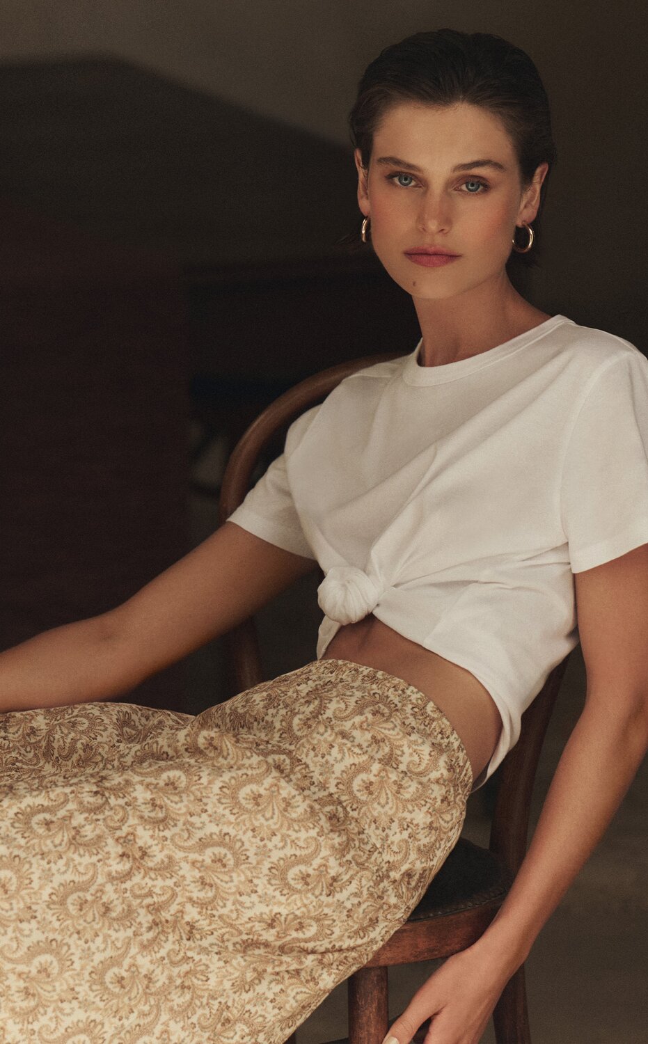 Woman wearing a white tee and a patterned midi-length skirt, lounging on a chair in an outdoor area.