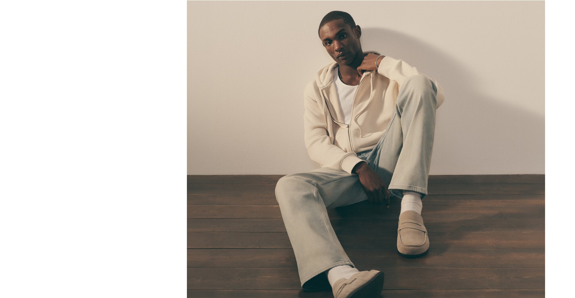 A man sitting on the floor wearing a white t-shirt, a tan zip up hoodie, light blue denim jeans and tan loafers. 