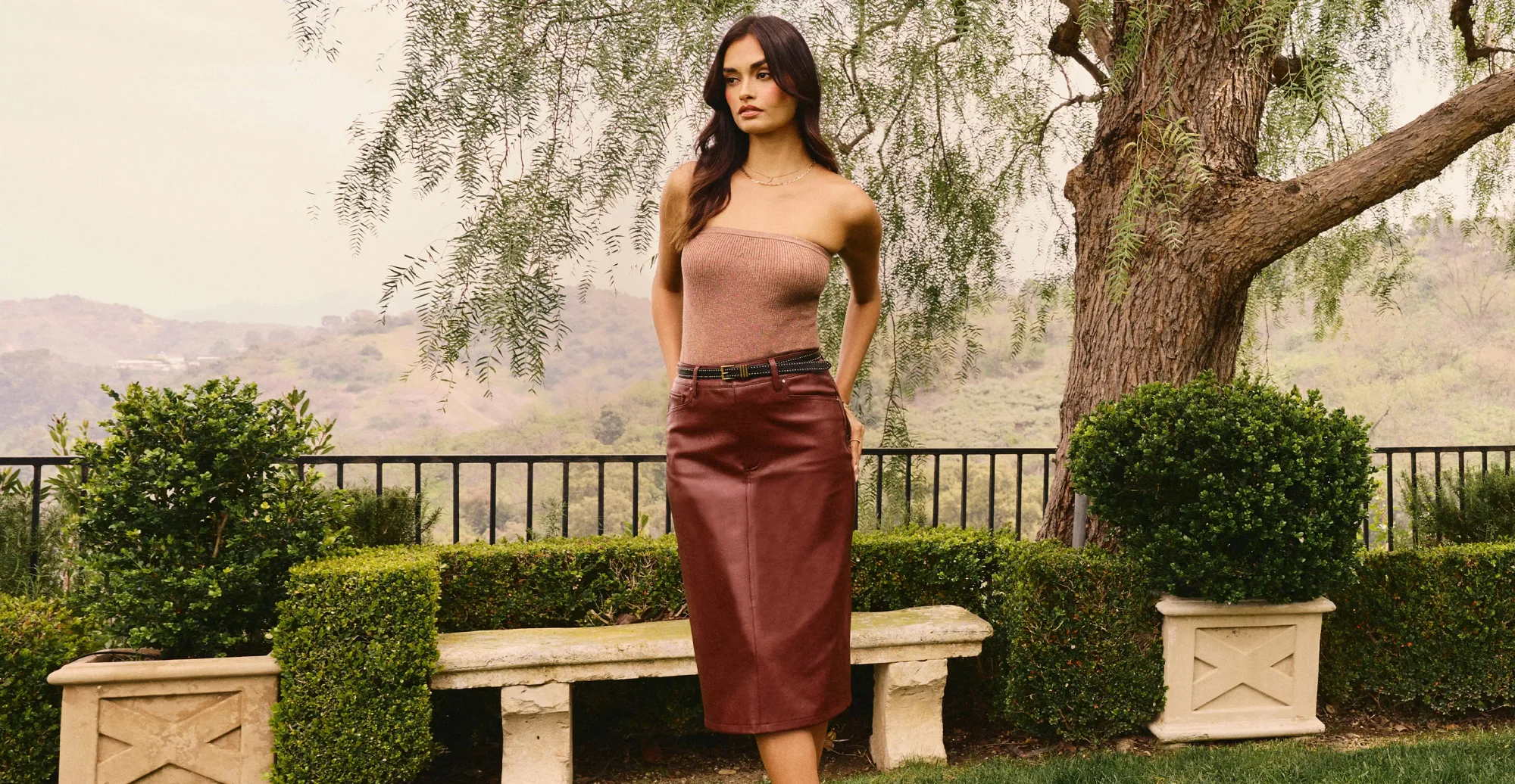 Woman standing outside wearing a brown metallic tube top, a black studded belt, and a dark brown faux leather midi skirt