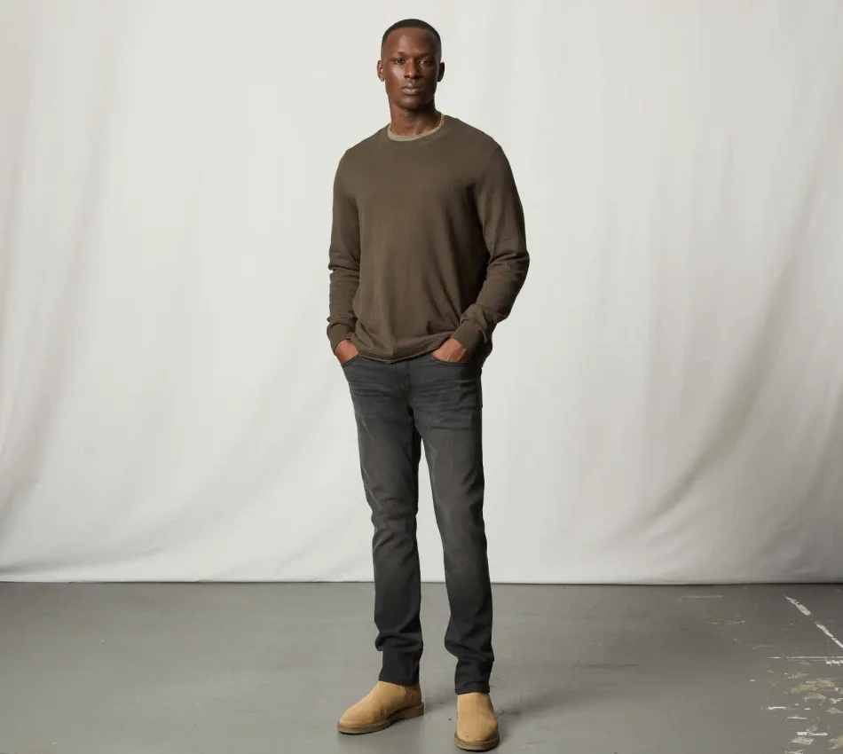 Man standing wearing a brown crewneck sweater, dark wash jeans, and tan suede boots