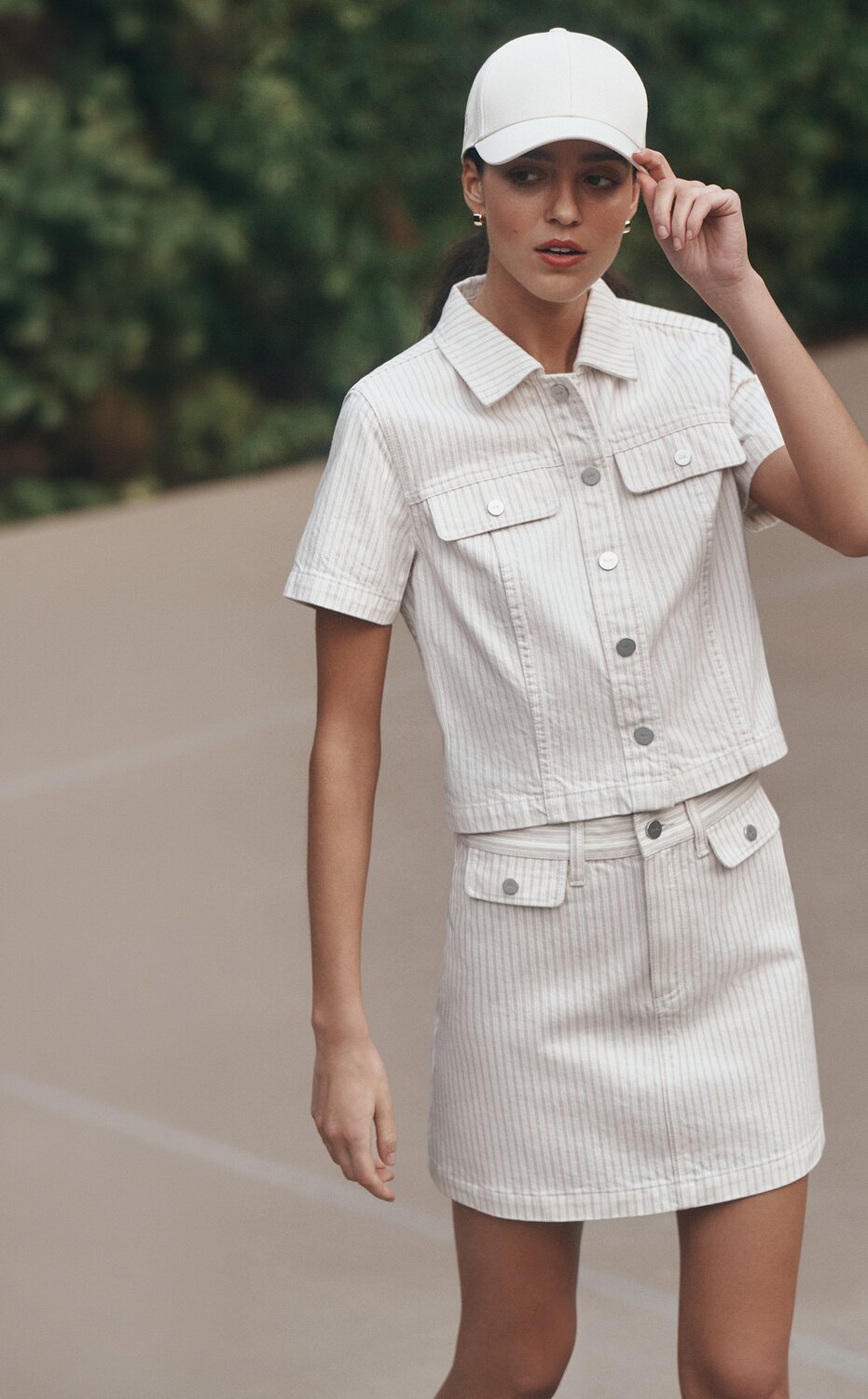 Woman posed at a tennis court wearing a white striped denim matching top and skirt with a white cap.