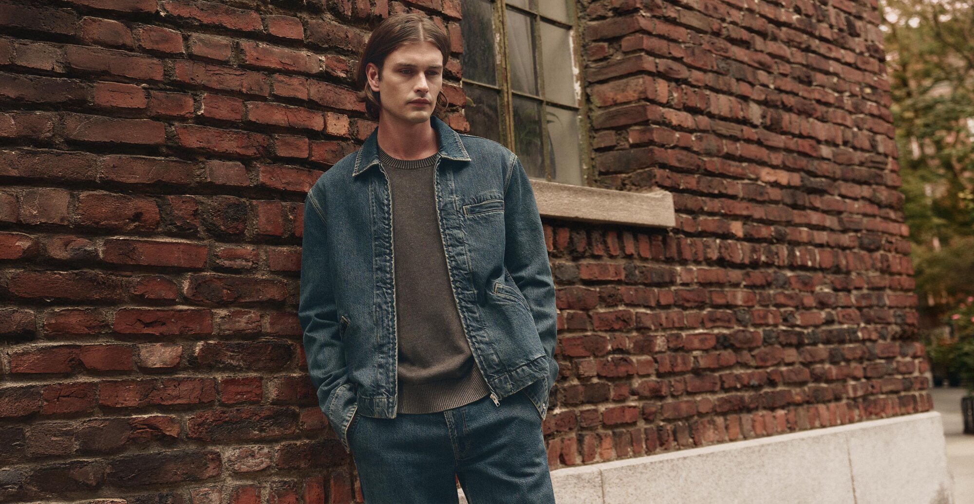 Man wearing a medium wash denim zip up jacket over a dark brown sweater and medium wash jeans