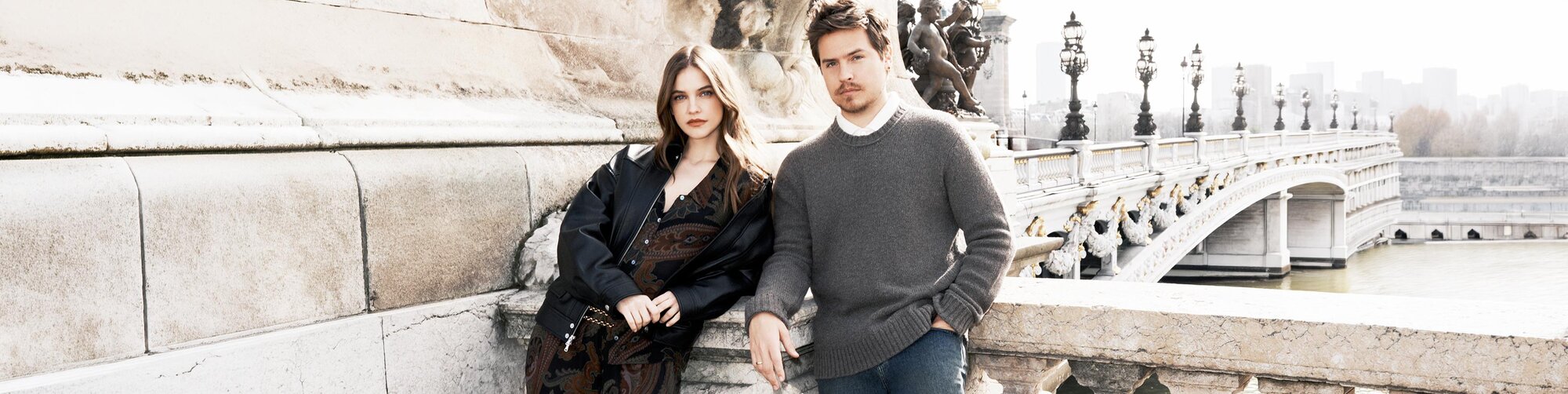 An image of Barbara Palvin and Dylan Sprouse for the Fall 2026 PAIGE campaign. Barbara wears a maroon and black paisley top and matching wide leg pants paired with a black oversized jacket in faux leather and gold chain belt. Dylan wears a grey sweater, white shirt and blue denim jeans.