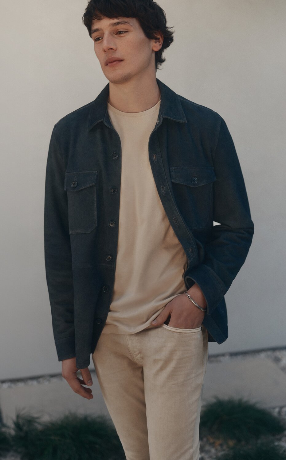 A man standing outside with his hand in his pocket wearing a navy blue suede jacket, a tan t-shirt, and tan denim jeans. 
