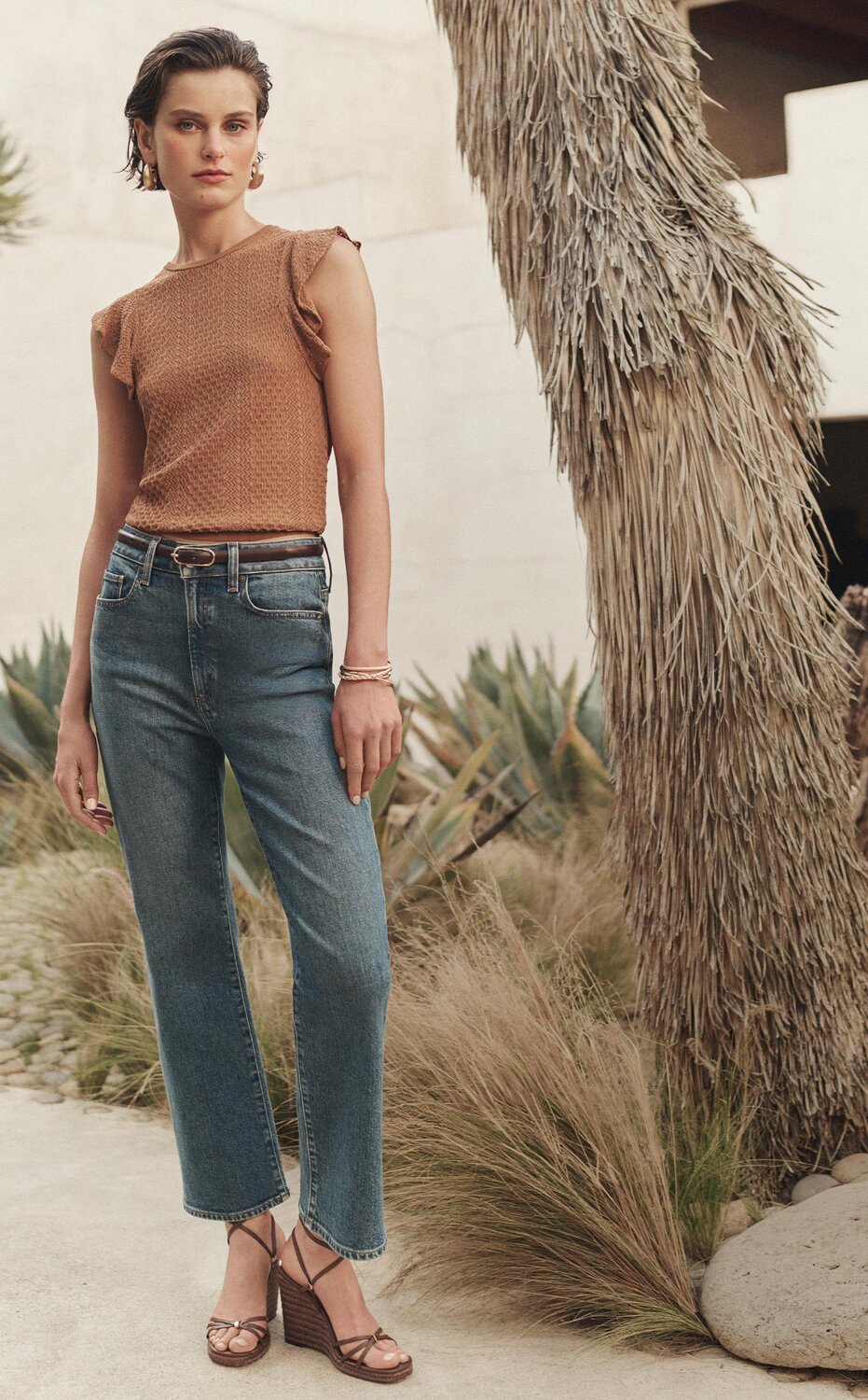 Woman wearing an orange flutter-sleeve knit top with a pair of medium wash high waisted straight-leg jeans and strappy brown wedge sandals, posed in a courtyard surrounded by palm trees.