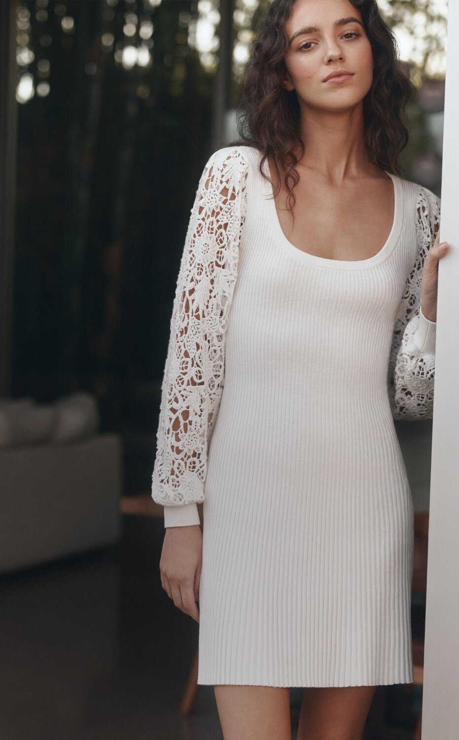 A woman standing in a doorway wearing a white knit mini dress with long floral embroidered sleeves