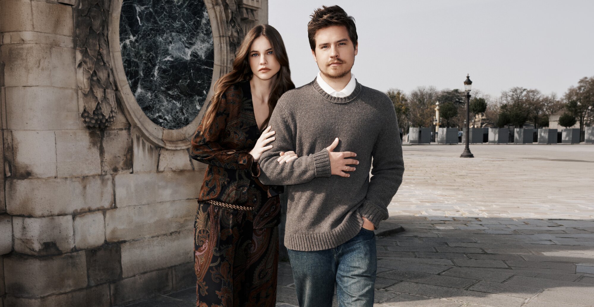Barbara Palvin wears a dark brown paisley print top and pant set with a gold chain belt as she walks arm-in-arm with Dylan Sprouse, who wears a grey crew neck sweater over a white button-up and a pair of medium wash jeans.