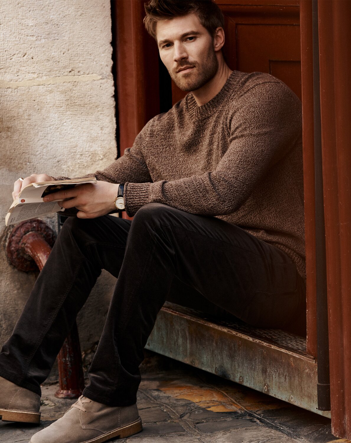 Male model wearing a light beige short-sleeve polo shirt, dark tailored shorts, white socks, and white sneakers, standing against a textured plaster wall.
