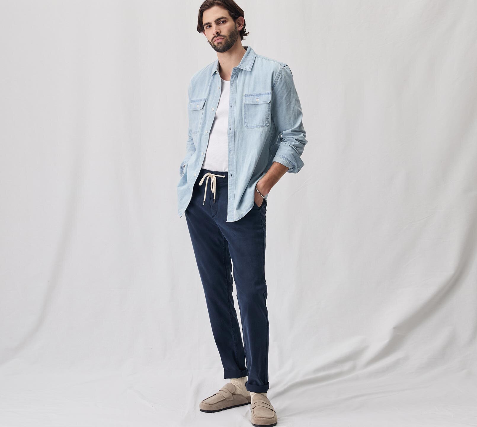 Man wearing a light wash denim button up shirt over a white tank and navy blue jogger pants