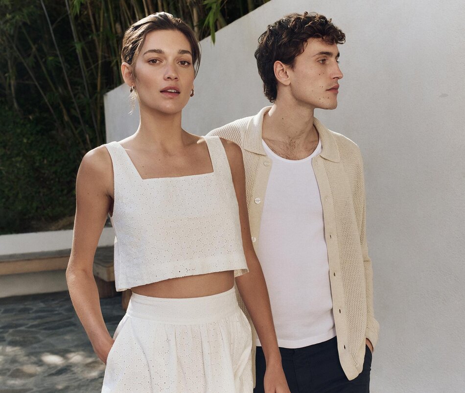 Woman wearing a white eyelet tank & skirt set & man wearing an ivory open knit button up, white tank & navy shorts