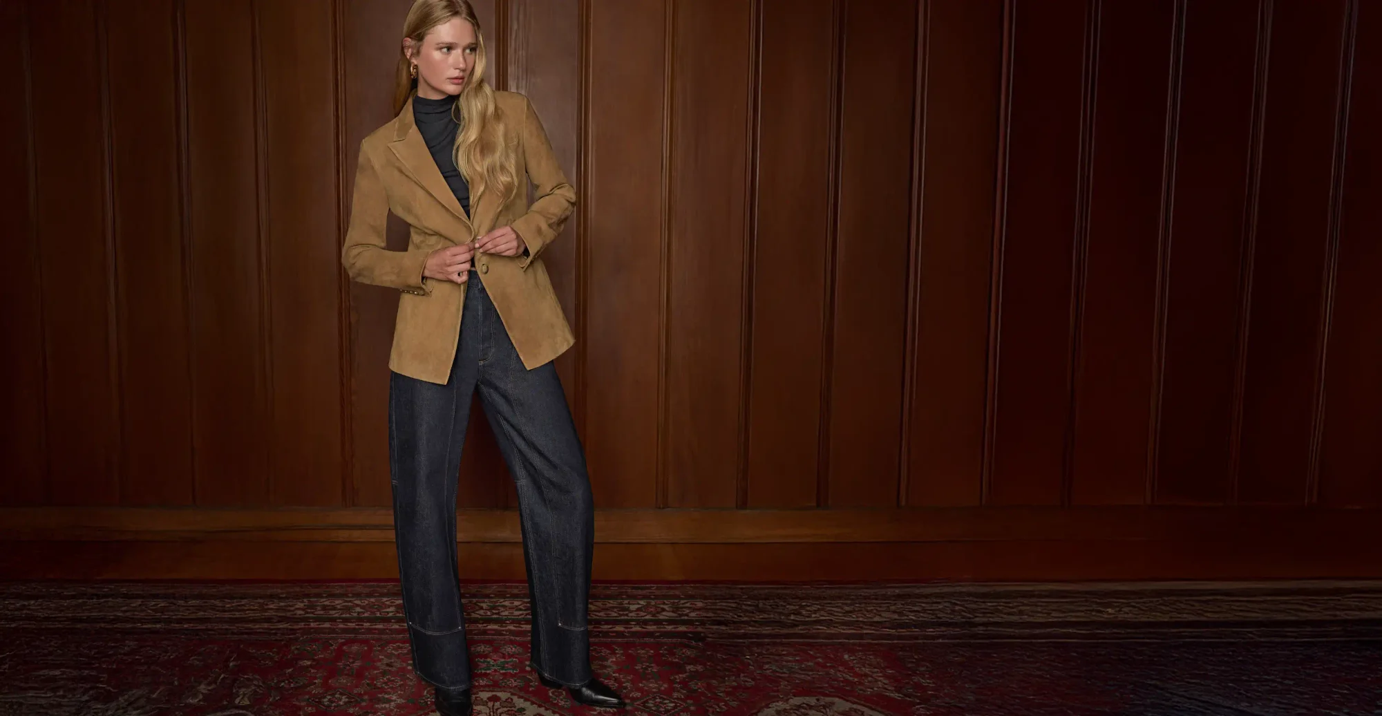 Woman standing wearing a tan suede blazer, a black top, and dark wash workwear jeans