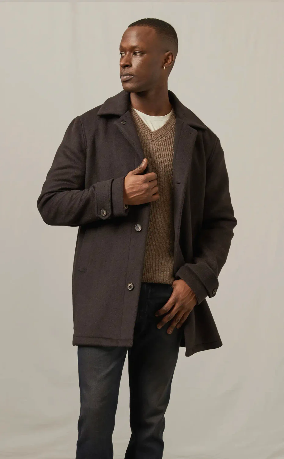 Man standing wearing a brown coat, brown sweater, and dark wash jeans